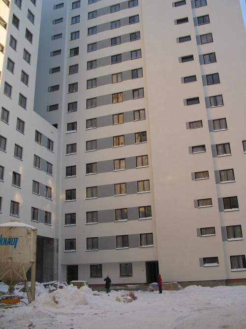 Wielopiętrowy budynek mieszkalny z oknami, widok z dołu na elewację z oknami, na pierwszym planie śnieg i dwie osoby, obok kontener Knauf.